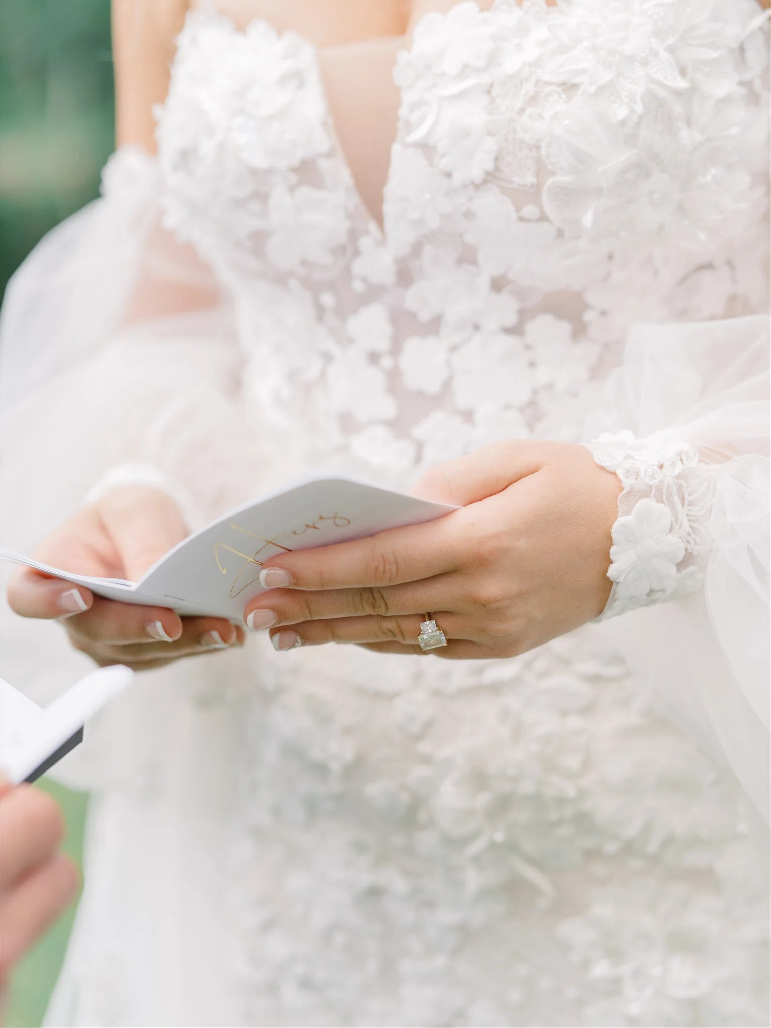 Close-up of a bride reading a wedding vow or speech, showing her wedding ring and intricate lace wedding dress with floral embellishments.