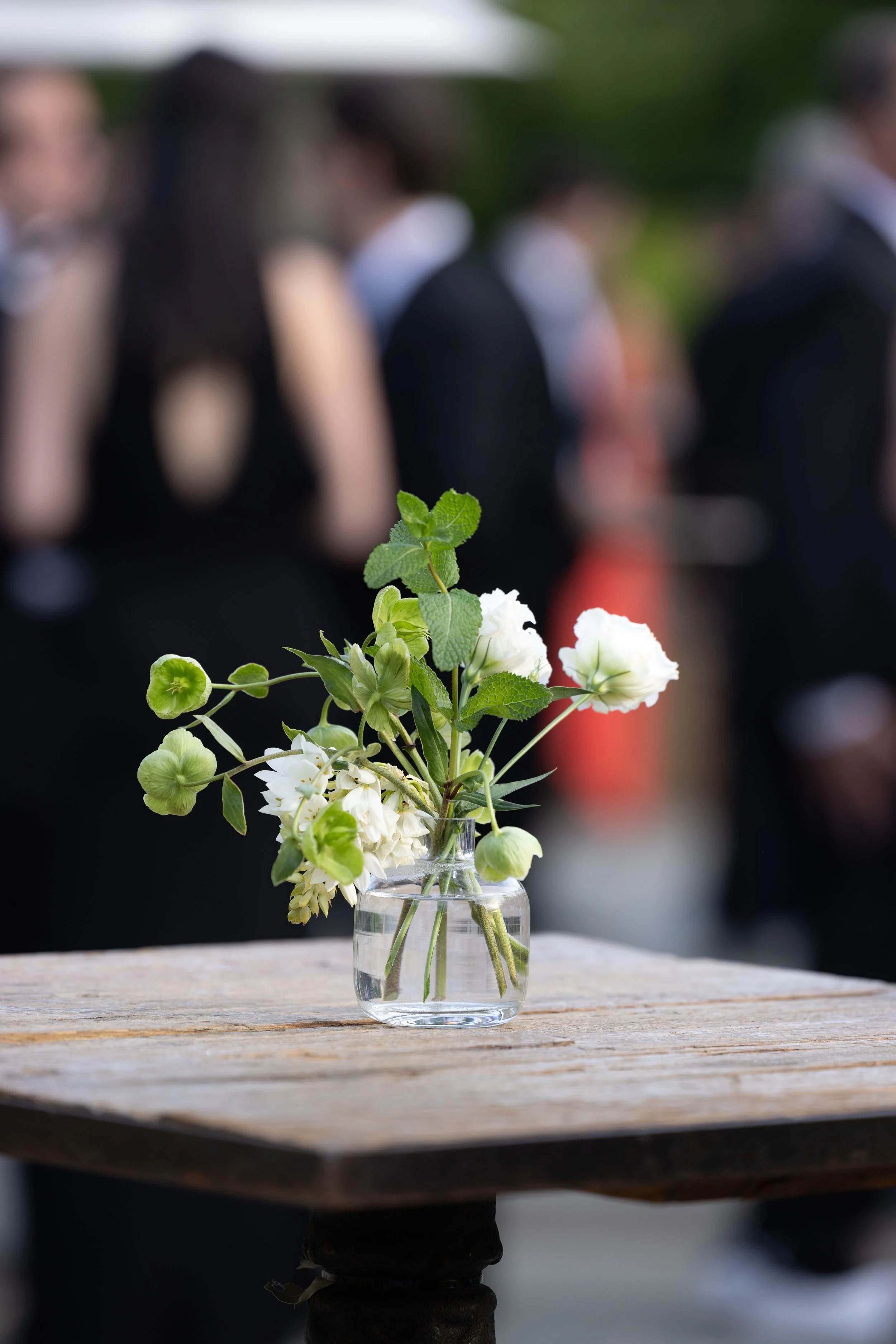 A small glass vase with white flowers and green leaves on a wooden table, blurred people in formal attire in the background.