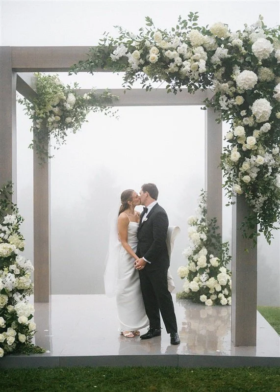 A bride and groom sharing a kiss under a decorated wedding arch adorned with white flowers and greenery.
