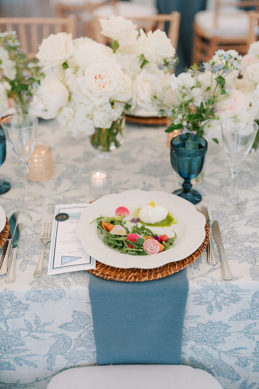 Elegant table setting with a white plate of salad, surrounded by silverware, glasses, and floral centerpieces with white and blush flowers, and a blue napkin underneath the plate.