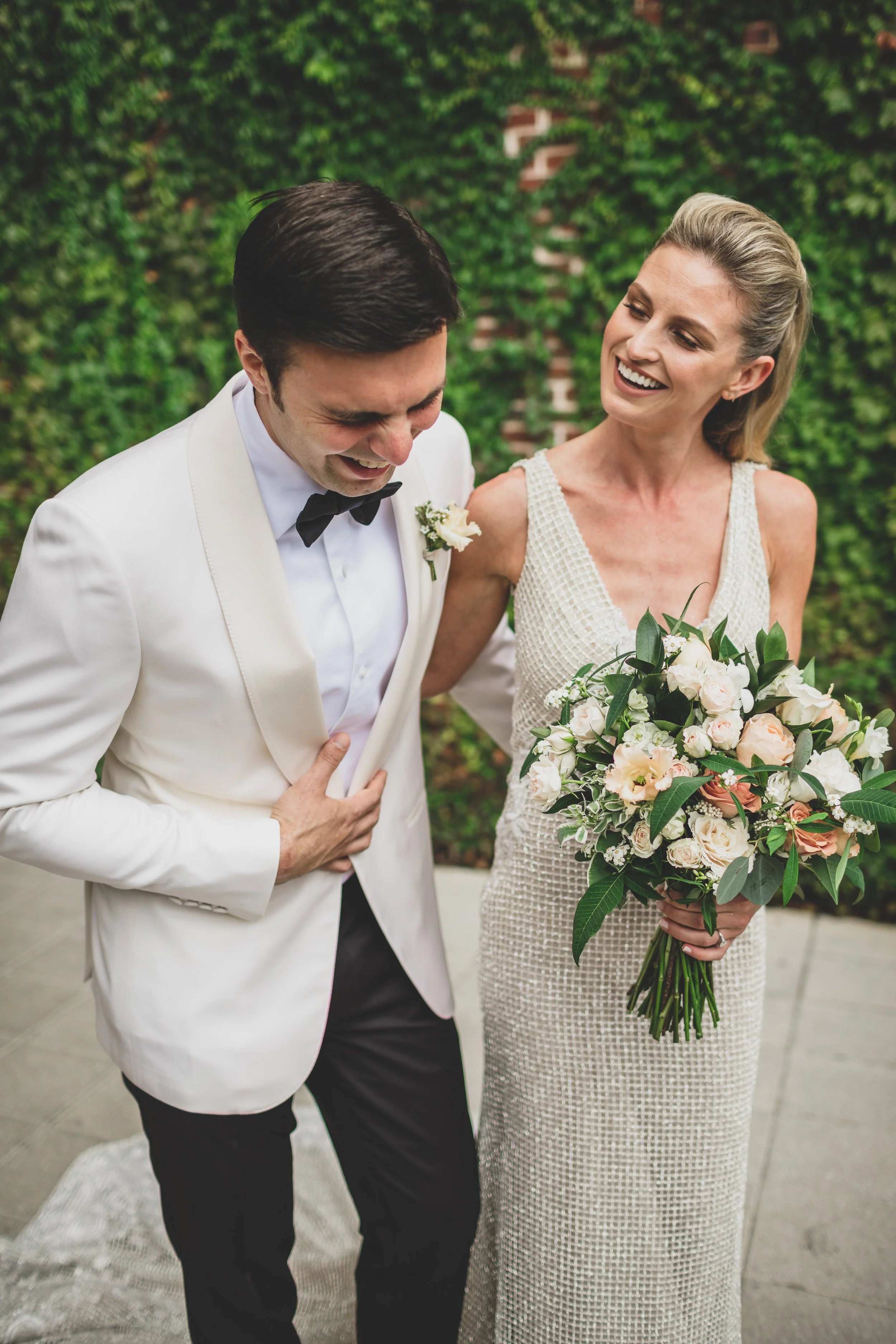 A bride and groom are laughing and sharing a joyful moment during their wedding. The groom is wearing a white tuxedo jacket, black pants, and a black bow tie, and is holding his stomach while looking down. The bride is wearing a sleeveless, textured,