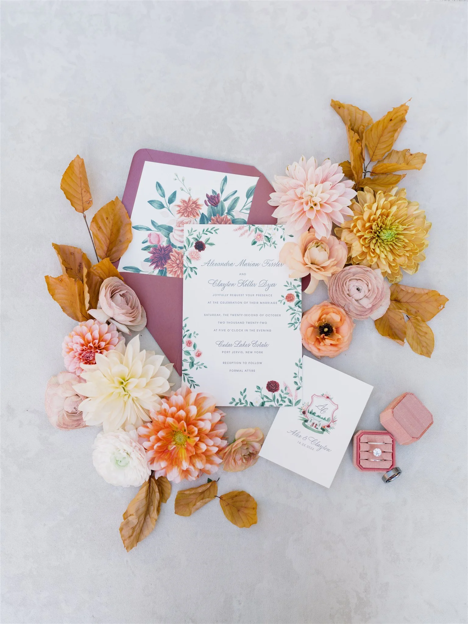 Wedding invitation surrounded by pastel-colored flowers and brown leaves, with a jewelry box containing a ring and a wedding band next to it.