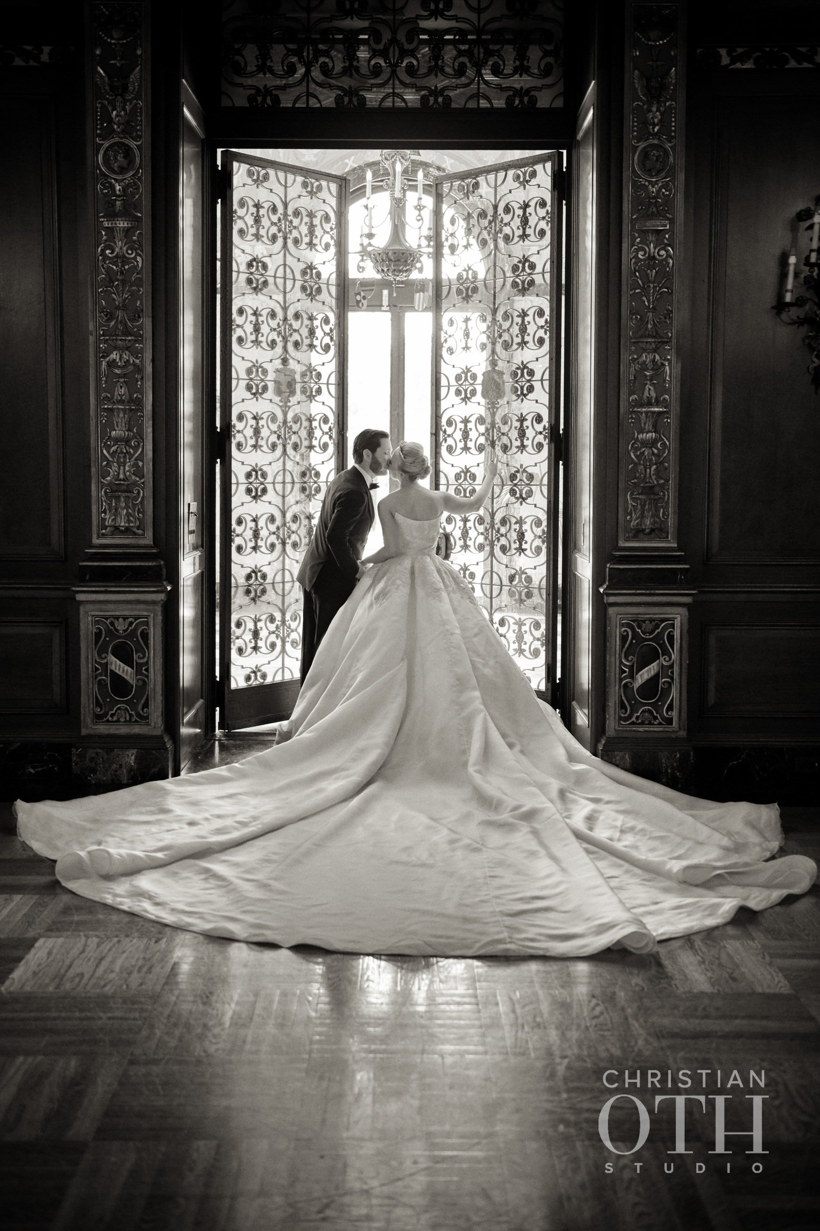 Black and white photo of a bride and groom standing near ornate doors with ironwork, inside a grand room with wooden flooring. The bride is in a voluminous gown with a long train, and the groom is in a suit. The bride is touching the door with her ri