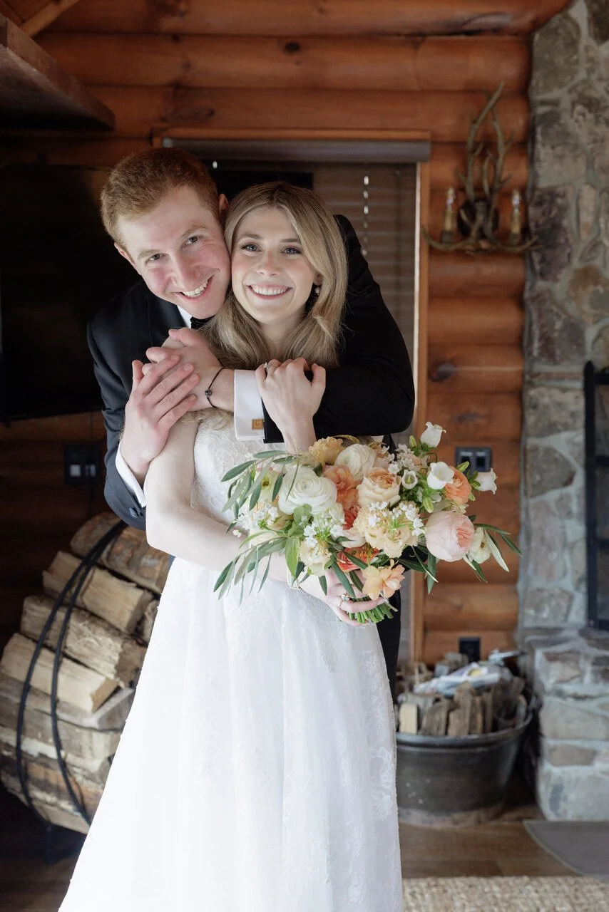 A newlywed couple smiling and hugging indoors, with the bride holding a bouquet of flowers.