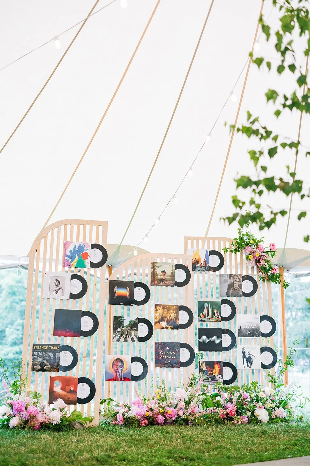 A decorated photo display board at an outdoor event, with photographs, vinyl record images, and floral arrangements at the base, under a white tent with string lights and greenery.