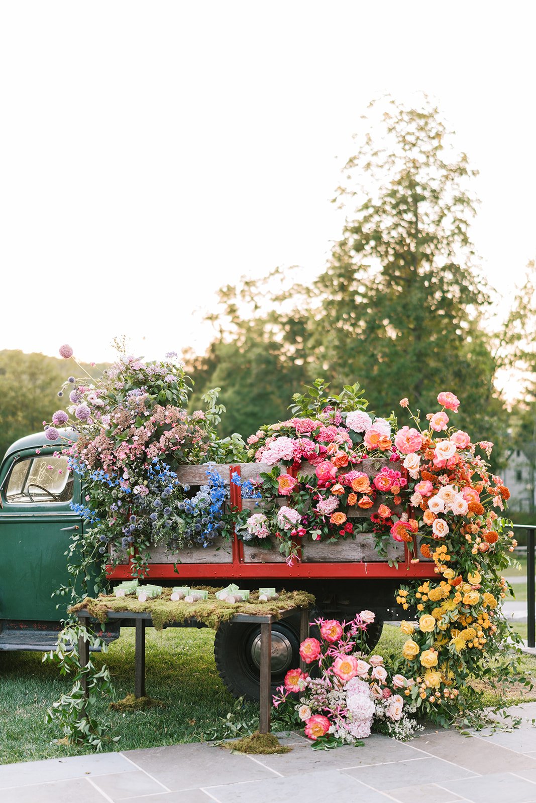 A vintage green truck with a red bed is filled with colorful flowers, including roses, hydrangeas, and other blooms, for a flower display or event.