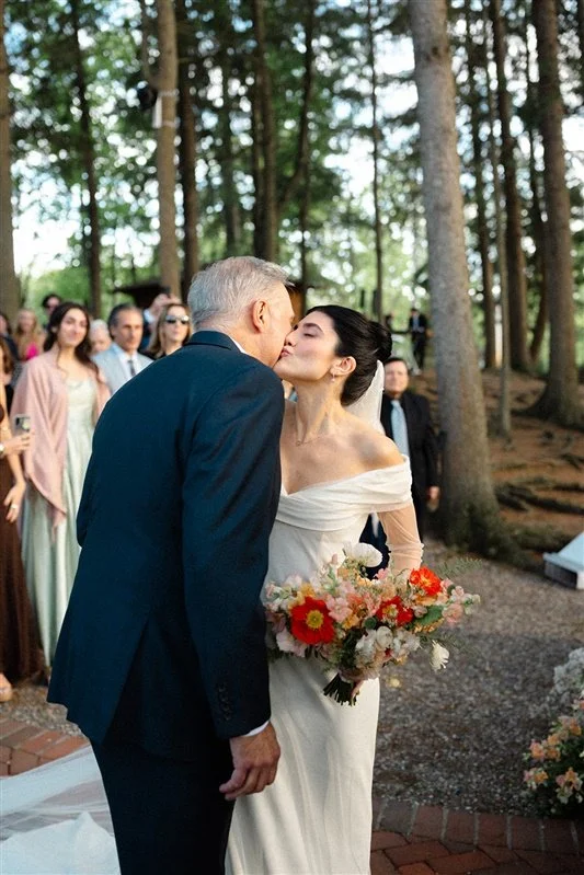 A bride and an older man are sharing a kiss at an outdoor wedding ceremony surrounded by trees. The bride holds a bouquet of colorful flowers, and guests are in the background.