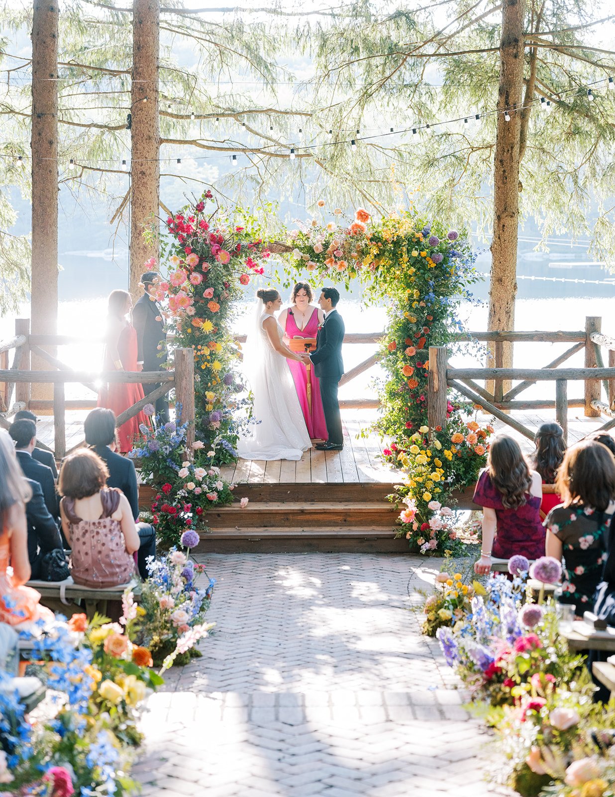 A wedding ceremony taking place on a wooden platform under tall pine trees by a lake, decorated with a colorful flower arch. The bride and groom are exchanging vows, surrounded by seated guests.