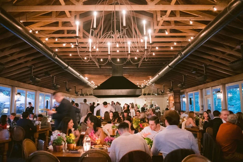A lively indoor restaurant with wooden ceiling beams, string lights, and large windows showing trees outside. Guests are seated at tables with flowers and candles, enjoying dinner. A server approaches a table in the foreground, and people are sociali