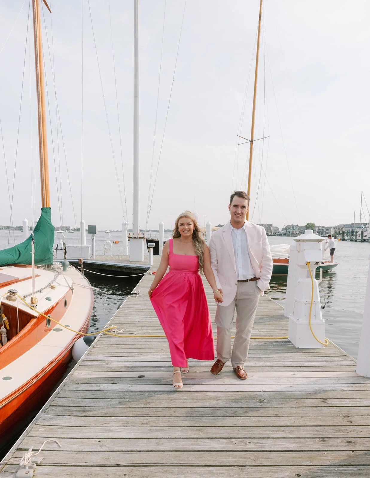A couple walking on a dock next to sailboats, holding hands, with a woman in a pink dress and a man in a white blazer and beige pants, by the waterfront.