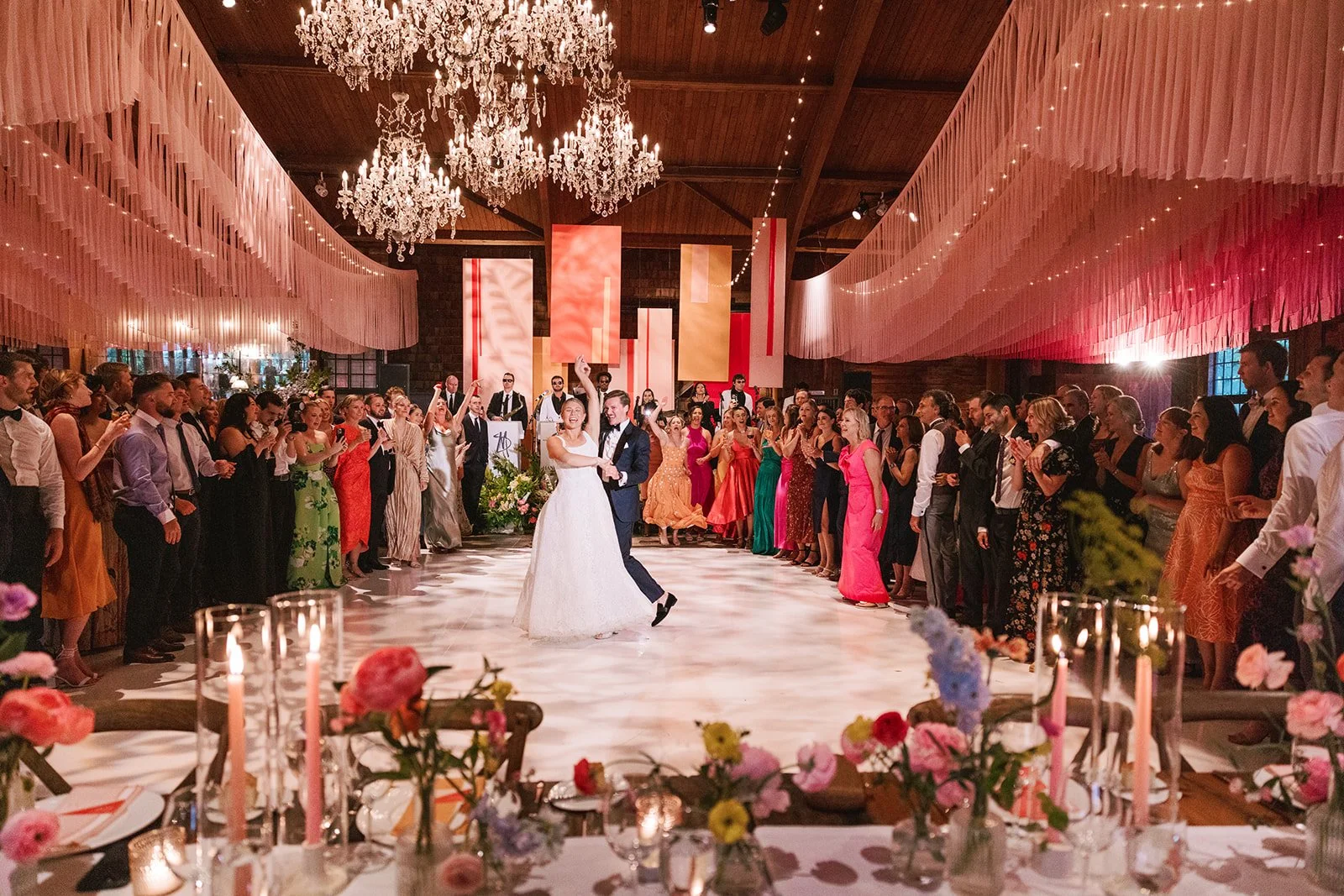 A bride and groom dance together in a large, elegant wedding reception hall filled with guests. The hall is decorated with chandeliers, hanging pink fabric, and colorful banners. Guests are standing around the dance floor, watching and clapping.