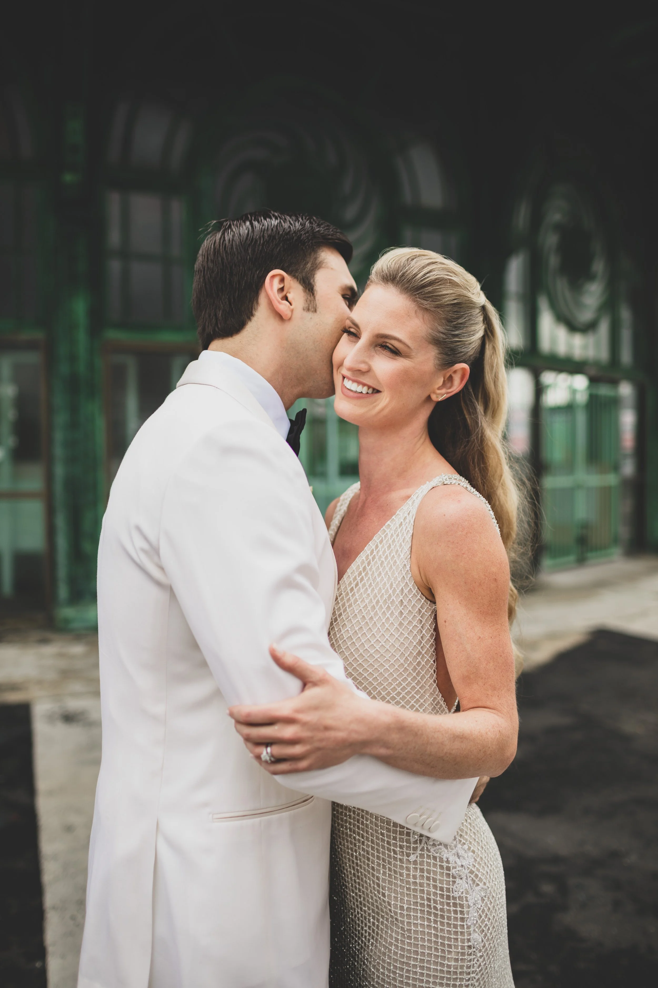 A couple dressed in wedding attire, smiling and embracing outside a building with green and black tinted windows.