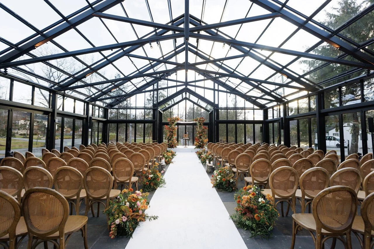 Indoor wedding ceremony setup with rows of wooden chairs on either side of a white aisle, decorated with colorful flowers, inside a glass greenhouse structure.
