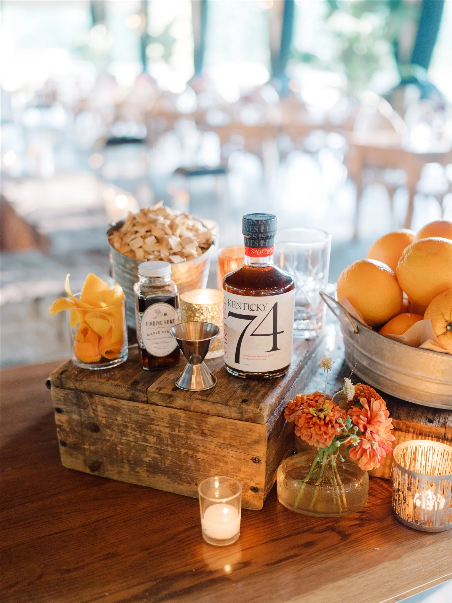 A rustic wooden table decorated with a jar of orange flowers, a lit tealight candle, a bottle of bourbon labeled Kentucky 74, a small bottle of maple syrup, a jigger, a glass, a metal bowl of lemons, and a large bowl of sliced fruits, set in a bright