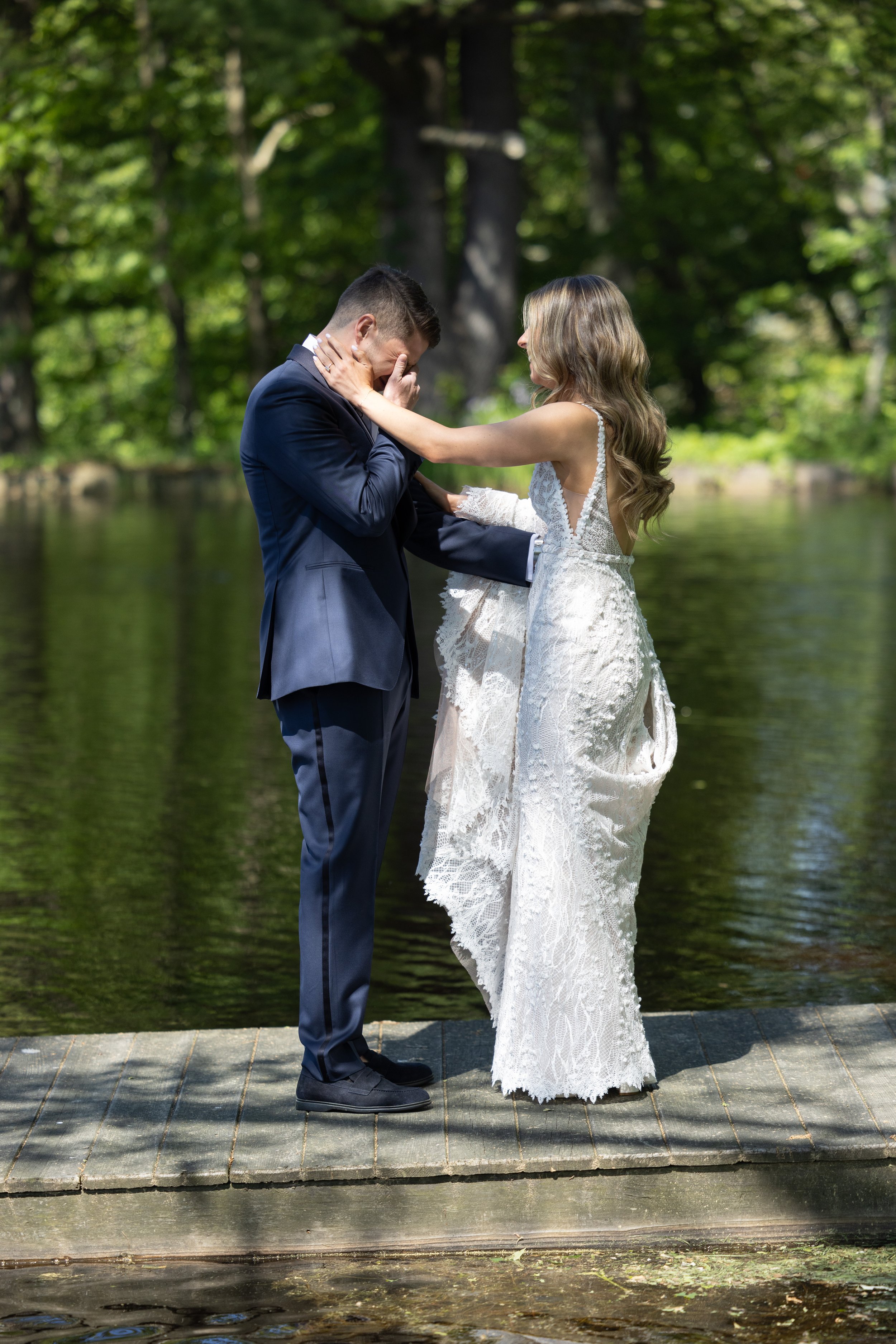 A bride and groom standing on a wooden platform by a river, with the bride touching the groom's face during their wedding ceremony in a forest setting.