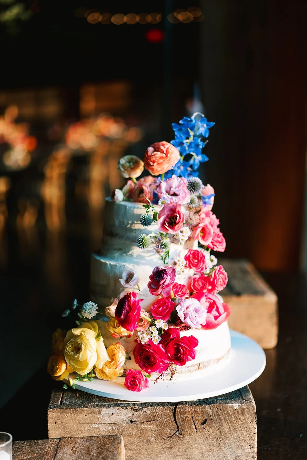 Three-tier wedding cake decorated with colorful flowers including roses and snapdragons, placed on a wooden table.