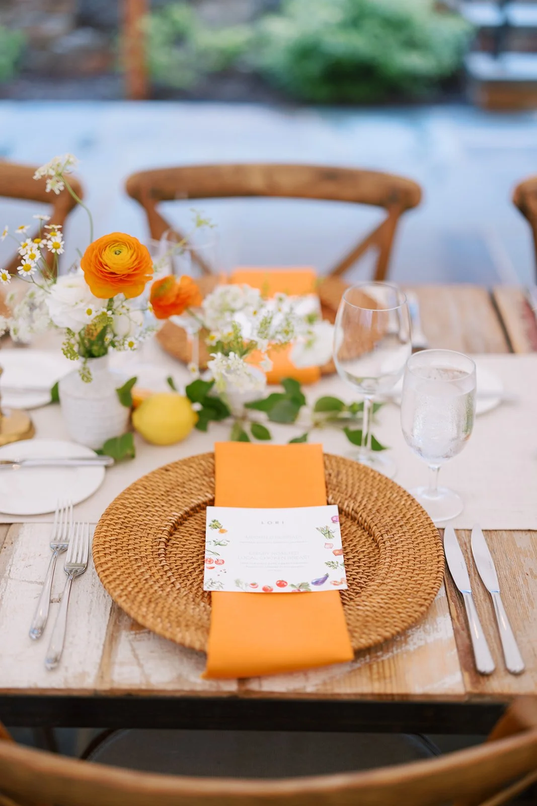 A rustic dining table set for a meal with a woven charger, orange napkin, and a printed menu. Centerpiece features white and orange flowers with greenery, a yellow lemon, and a lemon-shaped decorative object.