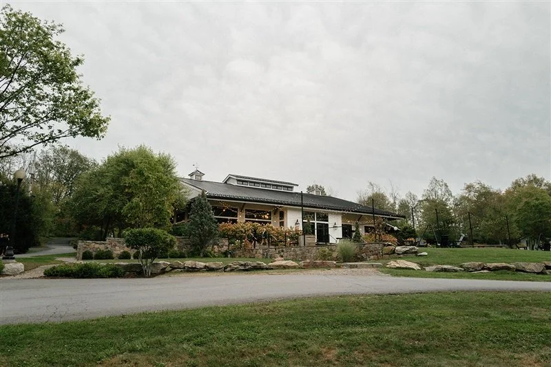 A large house with a stone and wood exterior, surrounded by trees and landscaped gardens, under an overcast sky.
