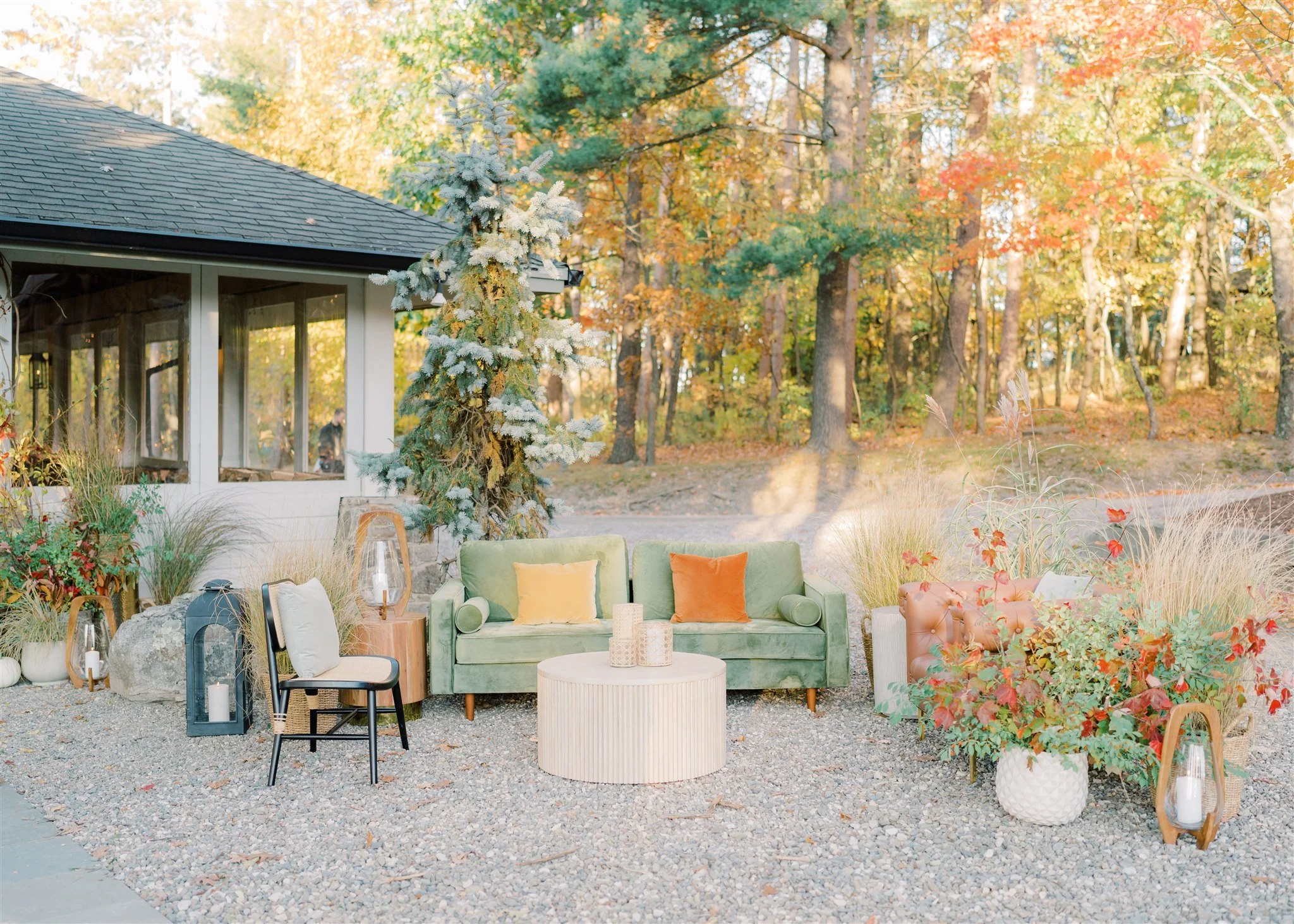 Outdoor living space with a green velvet sofa, a pink tufted armchair, and various decorative pillows and plants, set on a gravel ground amidst fall foliage with trees in the background.