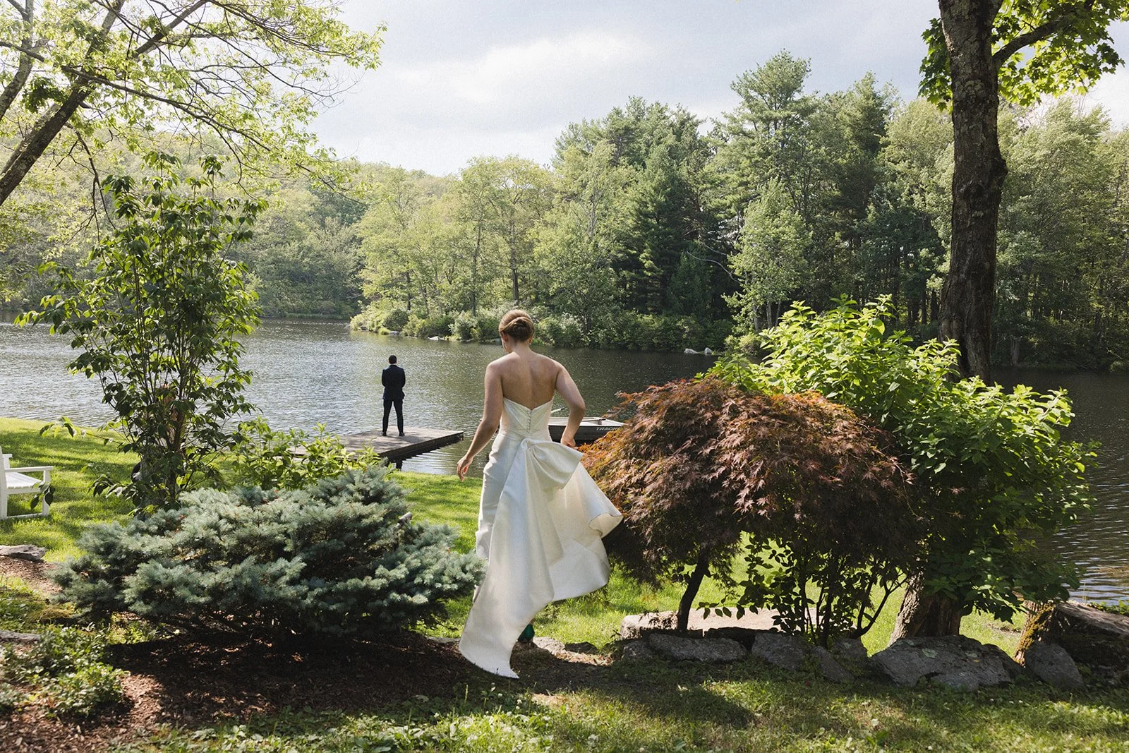 A person in a white wedding dress is standing on grassy area near a lake, looking at another person in a black suit standing on a dock by the water. The scene is surrounded by trees and lush greenery.
