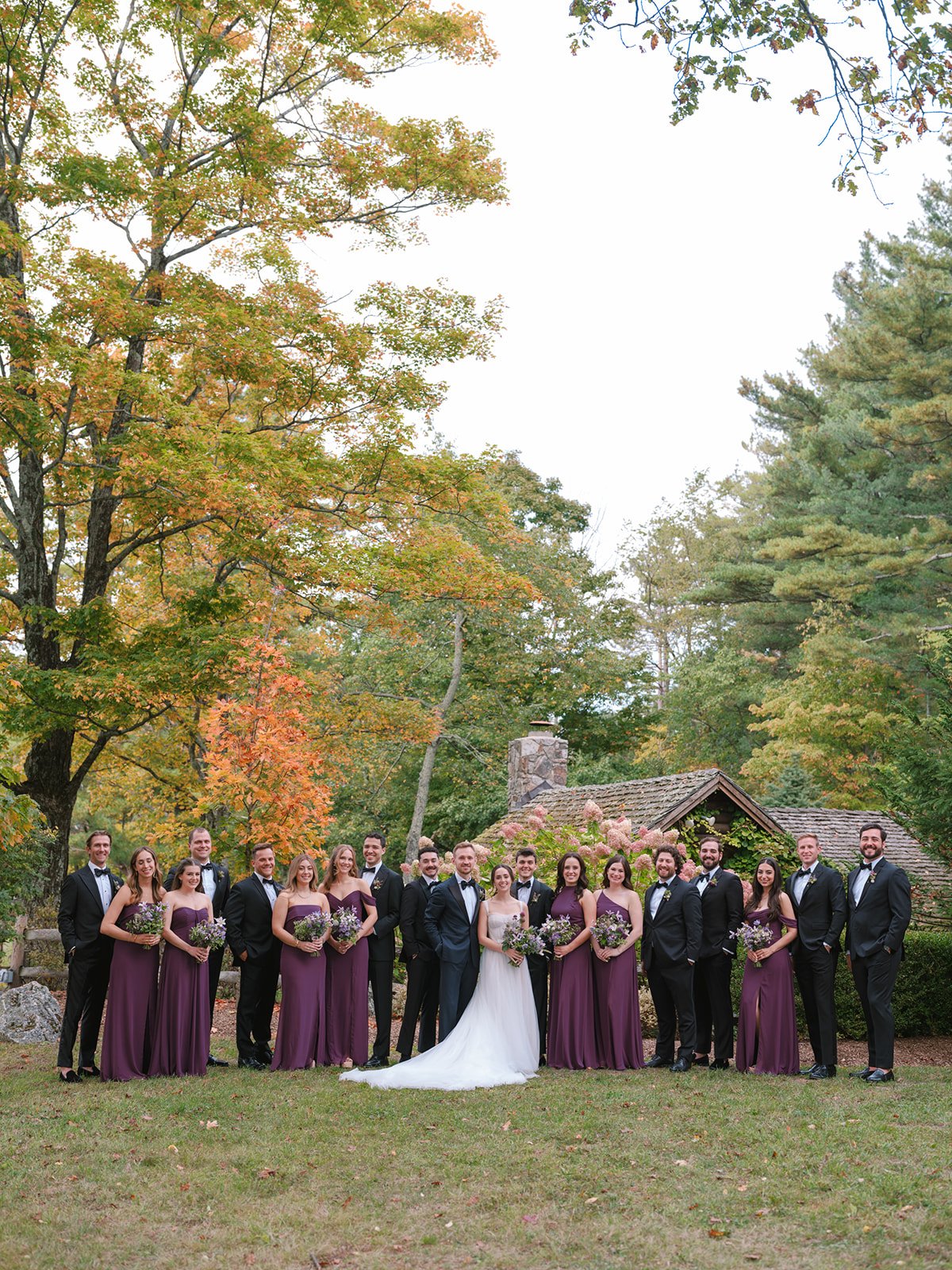 A wedding party with 16 members, including the bride in a white gown and the groom in a dark suit, standing outdoors in front of a rustic cottage, with trees showing autumn foliage.