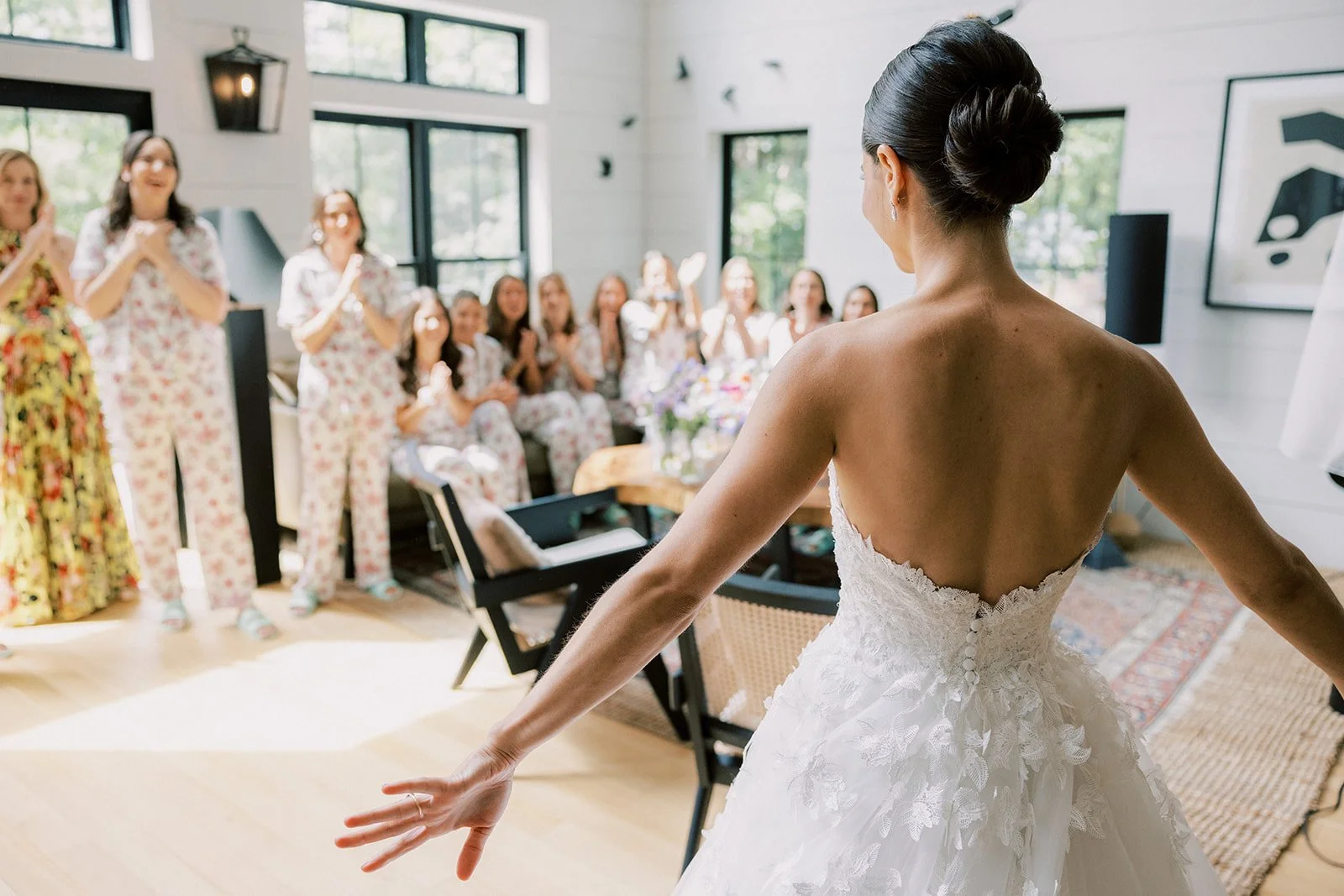 A bride in a white wedding dress with lace details standing in front of her bridesmaids, who are dressed in matching pajamas with floral patterns, inside a bright, modern room with large windows.