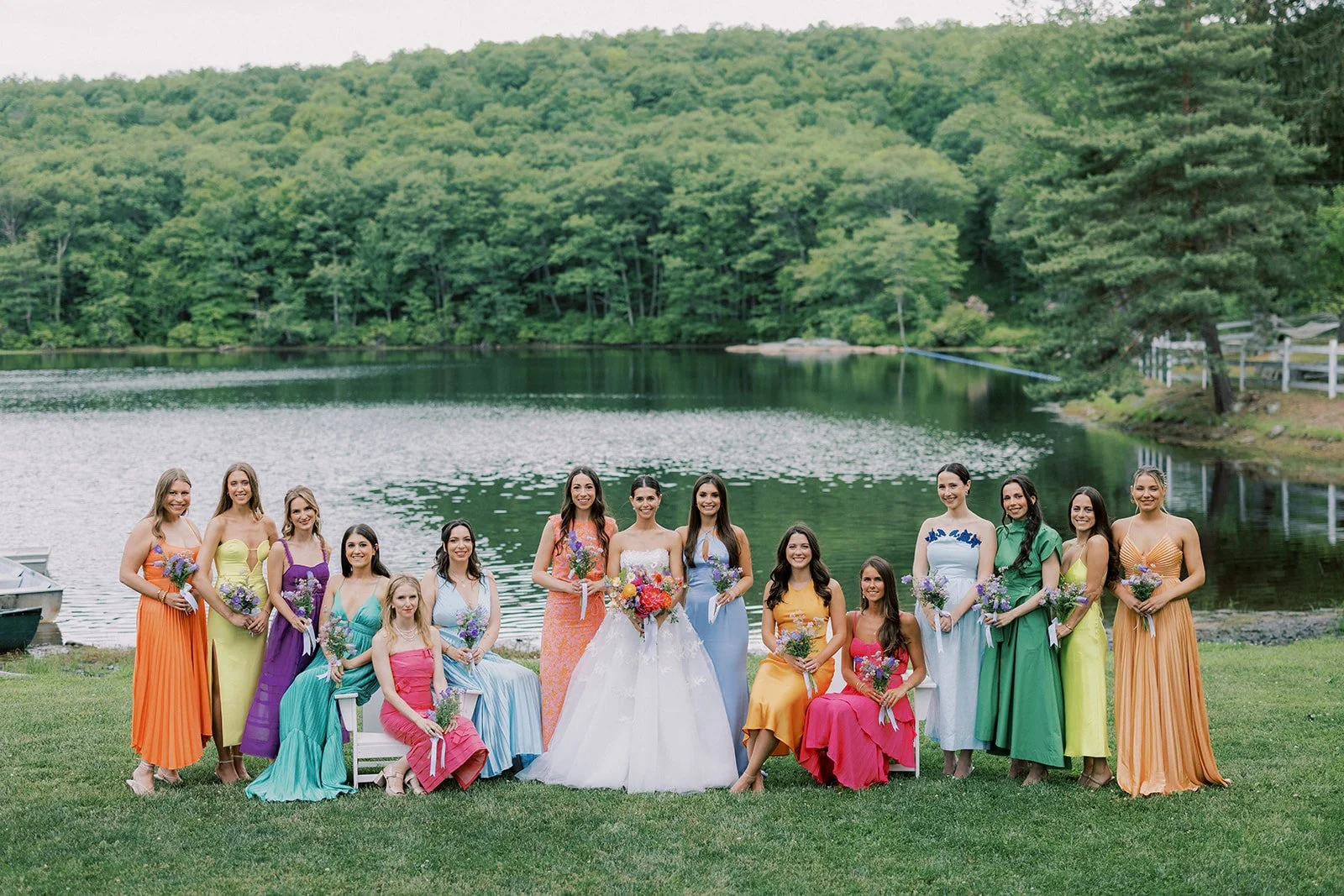 A group of women and girls in colorful dresses standing and sitting on a grassy area by a lake with a wooded hill in the background.