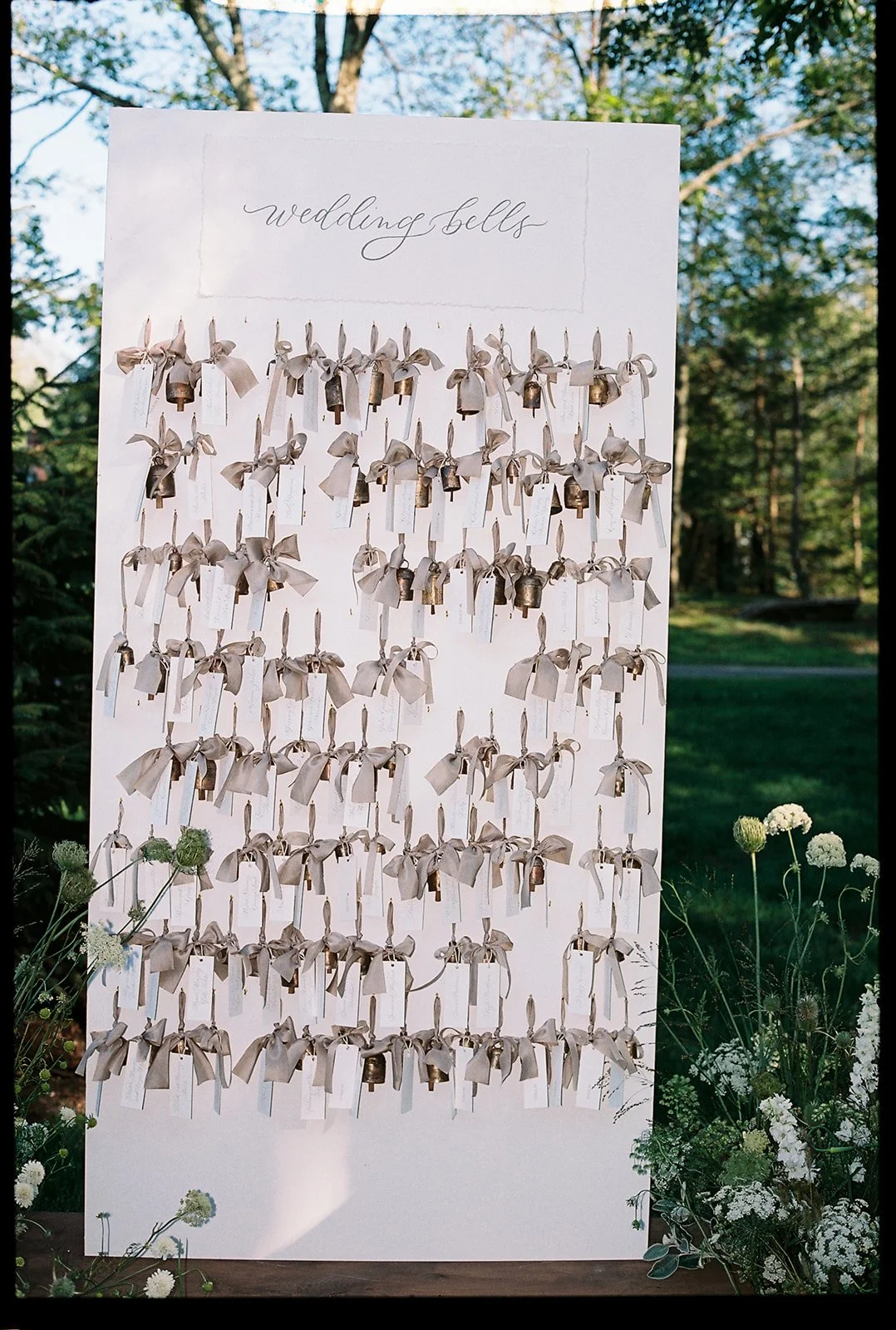 A white wedding bell table with numerous small bells hanging with beige ribbons, labeled as 'wedding bells', outdoors surrounded by green trees and white flowers.