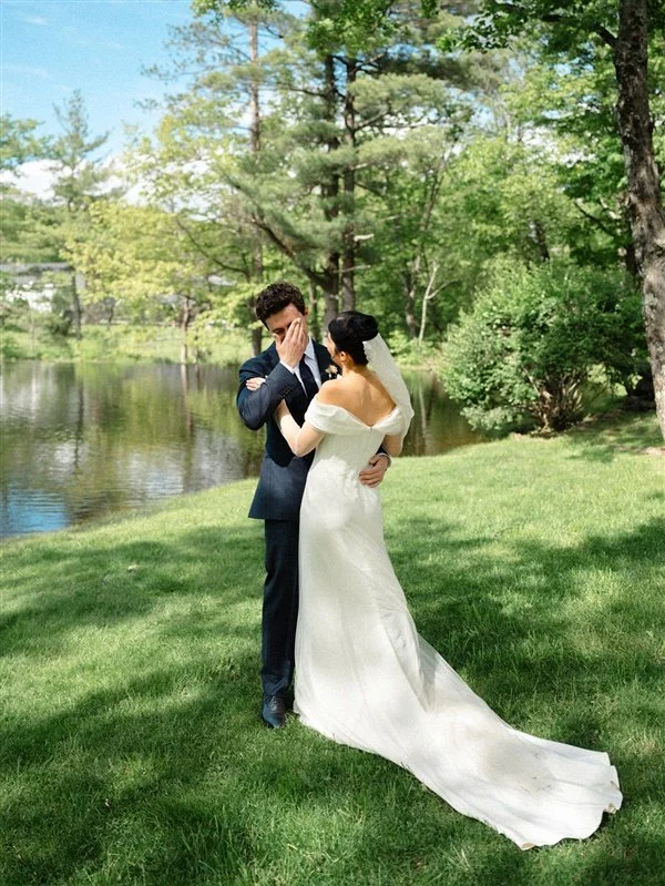 A newlywed couple stands by a lake in a green outdoor setting, with the groom covering his face and the bride holding his face gently.