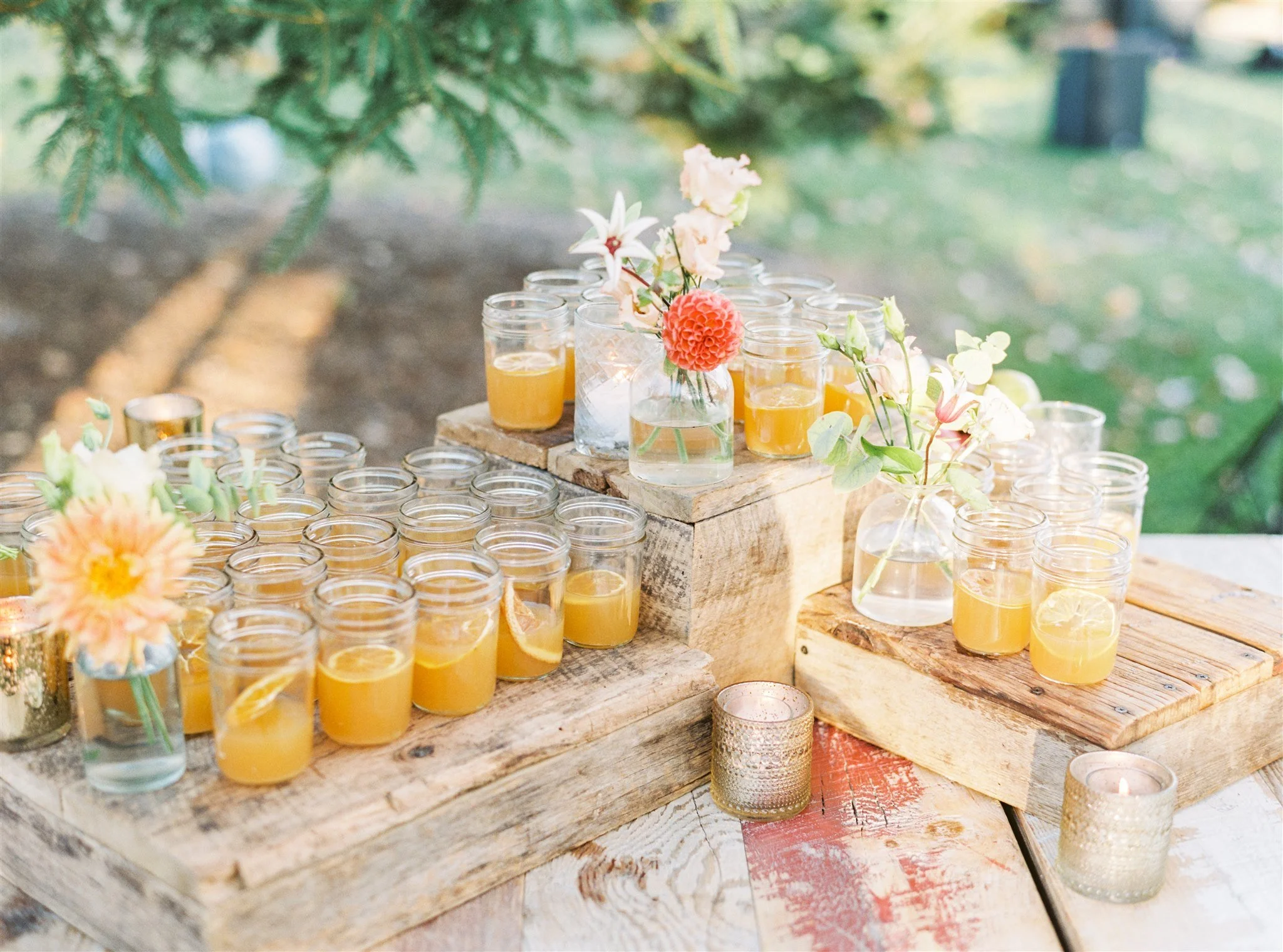 Glass jars with yellow drinks and floral arrangements on rustic wooden table outdoors.