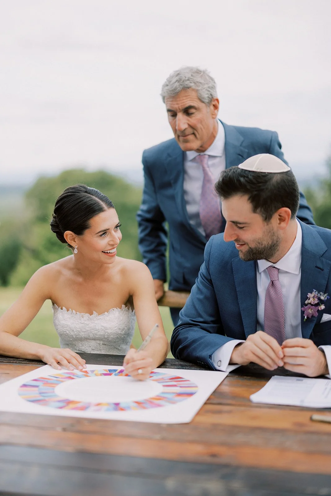 A bride and groom signing a document at an outdoor wedding, with an older man in a suit standing behind them, all smiling.
