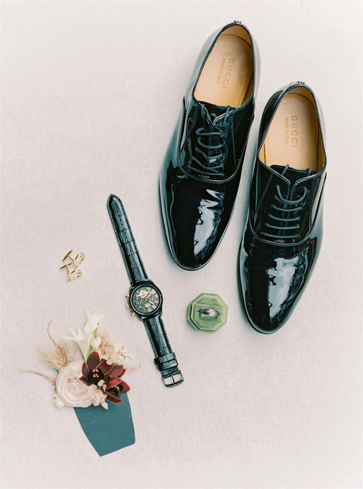 A pair of shiny black Gucci dress shoes, a black wristwatch with a leather strap, a gold ring with a gray stone, a gold ring with an inscription, a small boutonniere with white and burgundy flowers and greenery, and a small green box with a silver ri