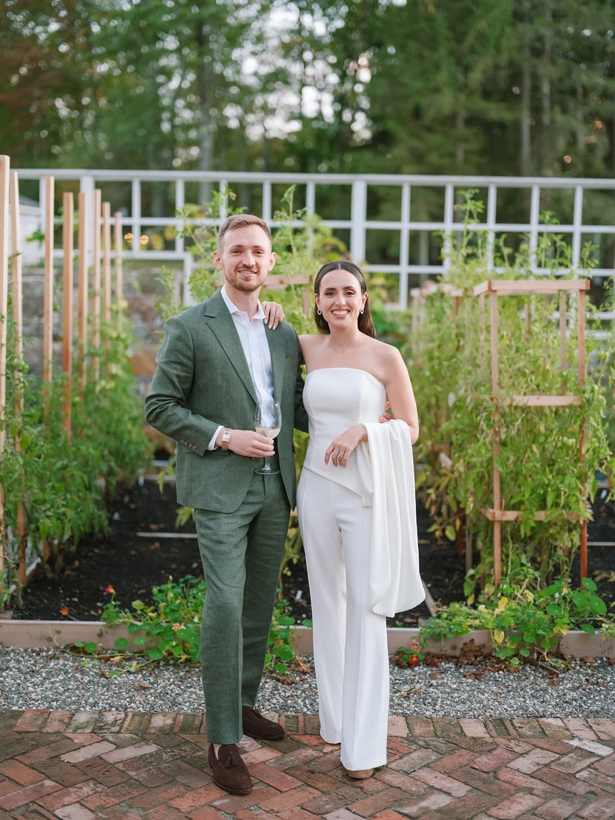 A couple dressed in formal attire standing outdoors on a brick path, holding glasses of wine, with a garden and greenery in the background.