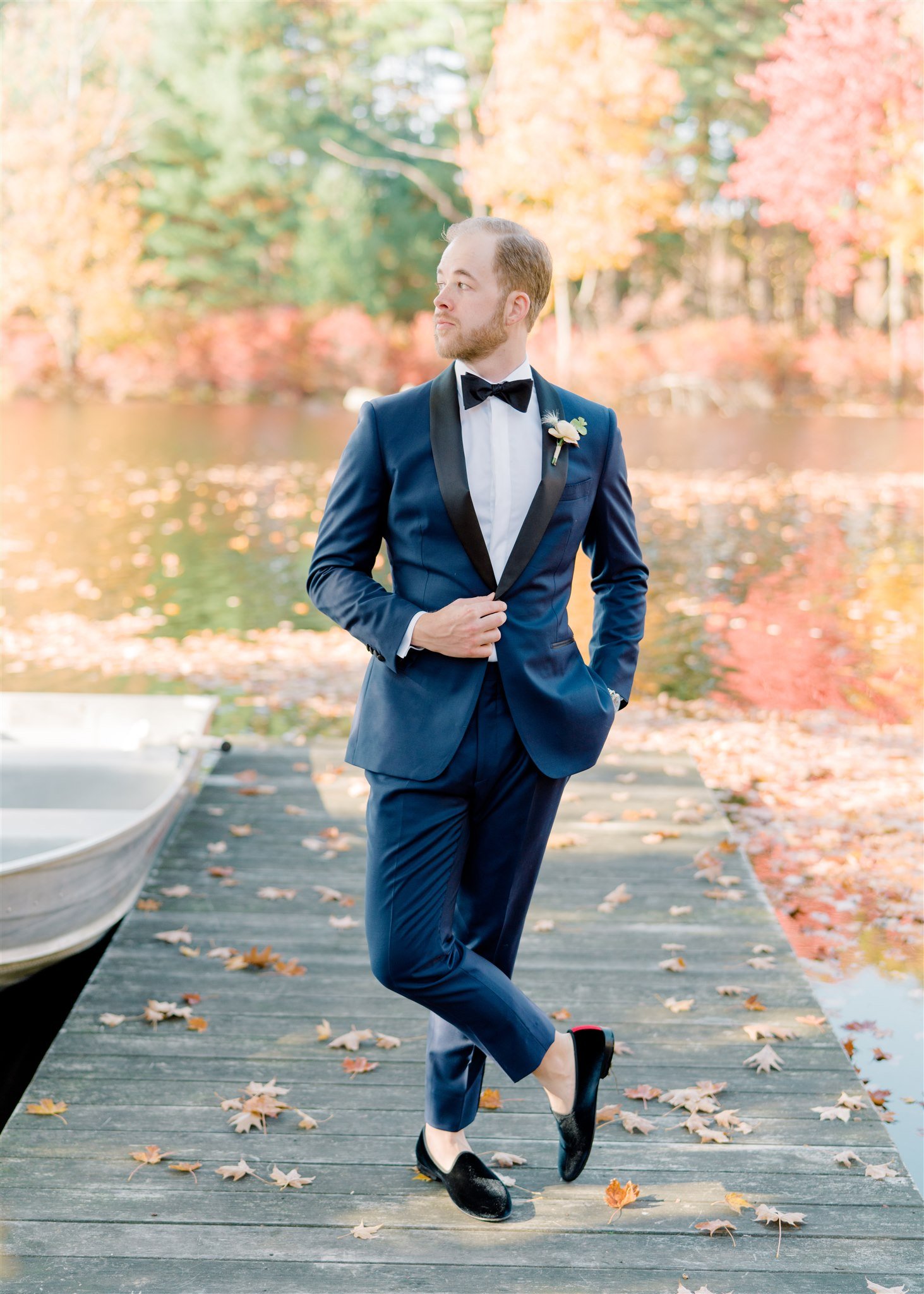 A man in a navy tuxedo with a black satin lapel and a bow tie is standing on a wooden dock near a lake. He has a boutonniere on his lapel and is looking to the side. Fall leaves are scattered on the dock, and autumn trees with orange, yellow, and red