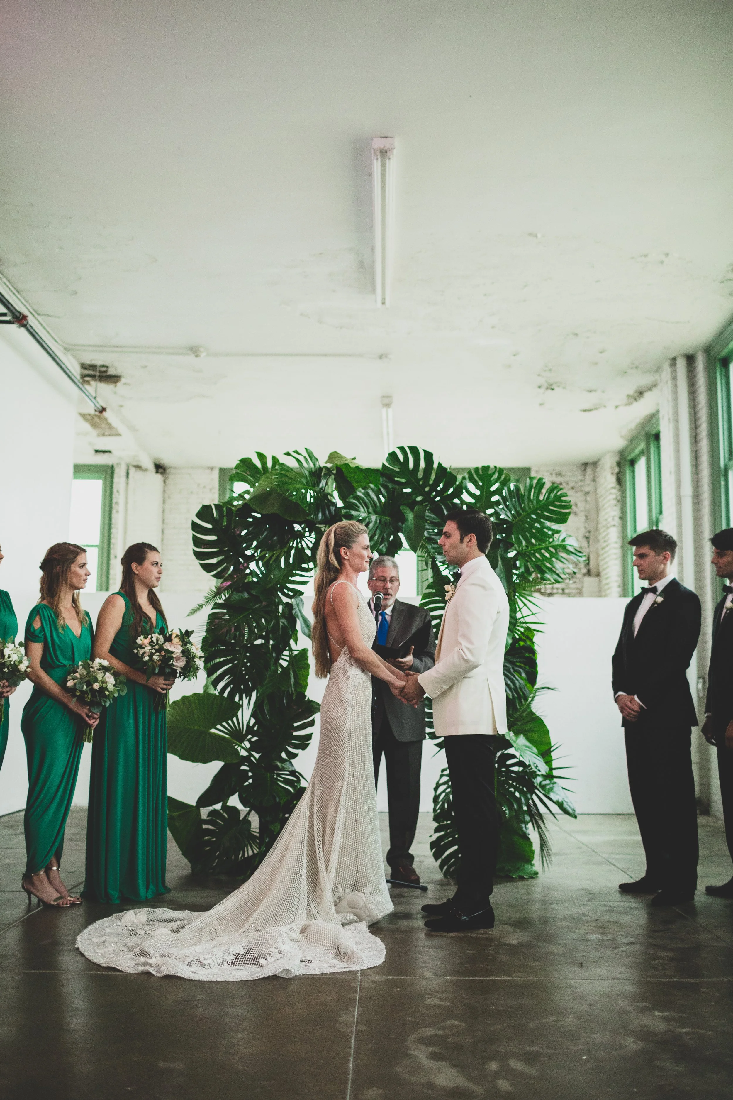 A wedding ceremony with a bride and groom holding hands, exchanging vows in front of an officiant in an industrial-style venue decorated with large tropical plants. Bridesmaids in teal dresses and groomsmen in black suits stand on either side, with l