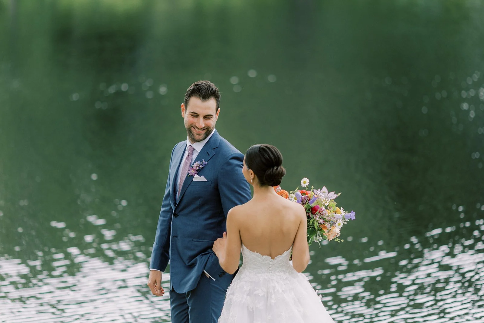 A bride and groom standing together near a body of water, with the groom smiling and looking at the bride. The bride is holding a bouquet and has her hair styled in an elegant bun.