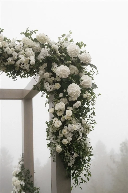 White floral wedding arch decorated with white roses, hydrangeas, and greenery, set outdoors on a foggy day.