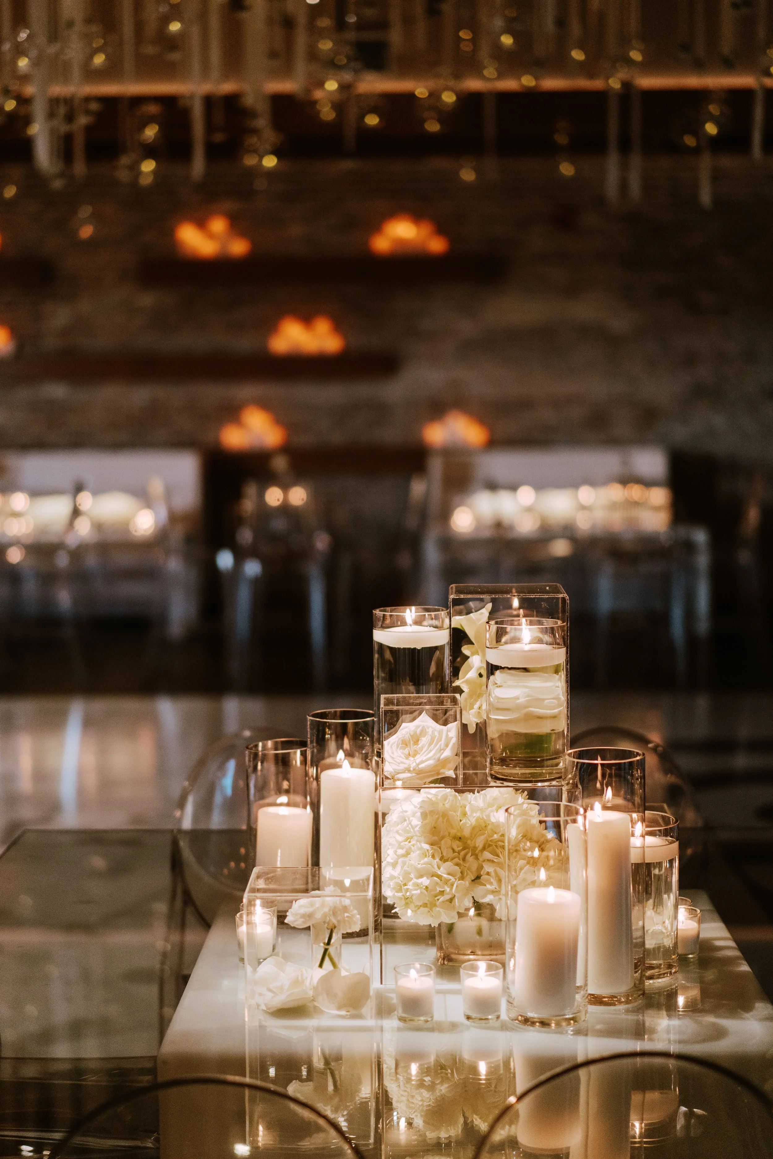 Elegant centerpiece with white flowers and tall candles on a table, set in a dimly lit venue with bokeh lights in the background.