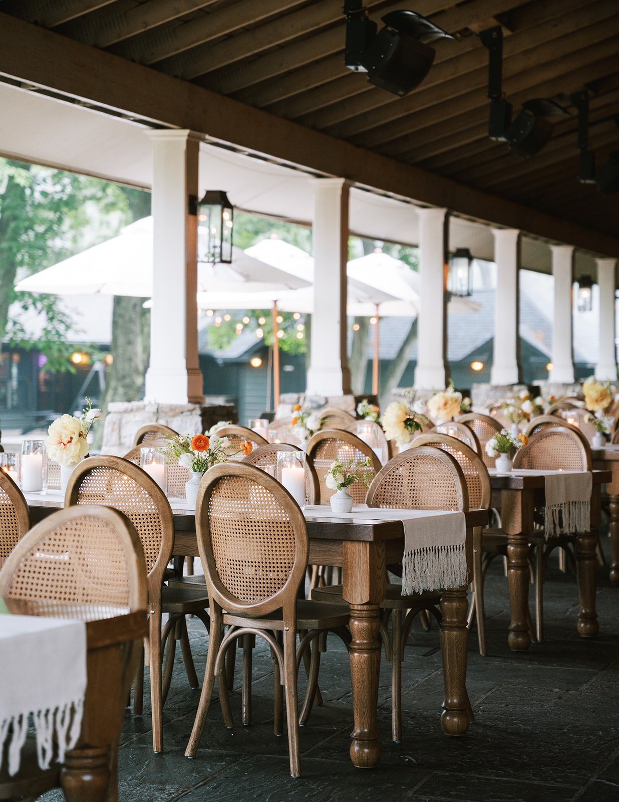 Wedding reception area with wooden tables and wicker chairs decorated with white tablecloths and floral centerpieces, outdoor patio with umbrellas and string lights.