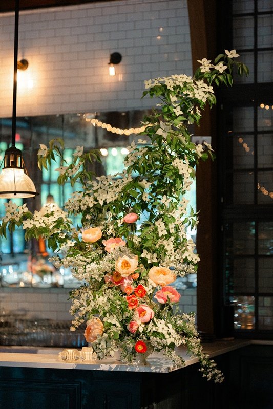 Large floral arrangement with white, pink, and red flowers and green foliage inside a restaurant or café, near windows with string lights and black metal frames.