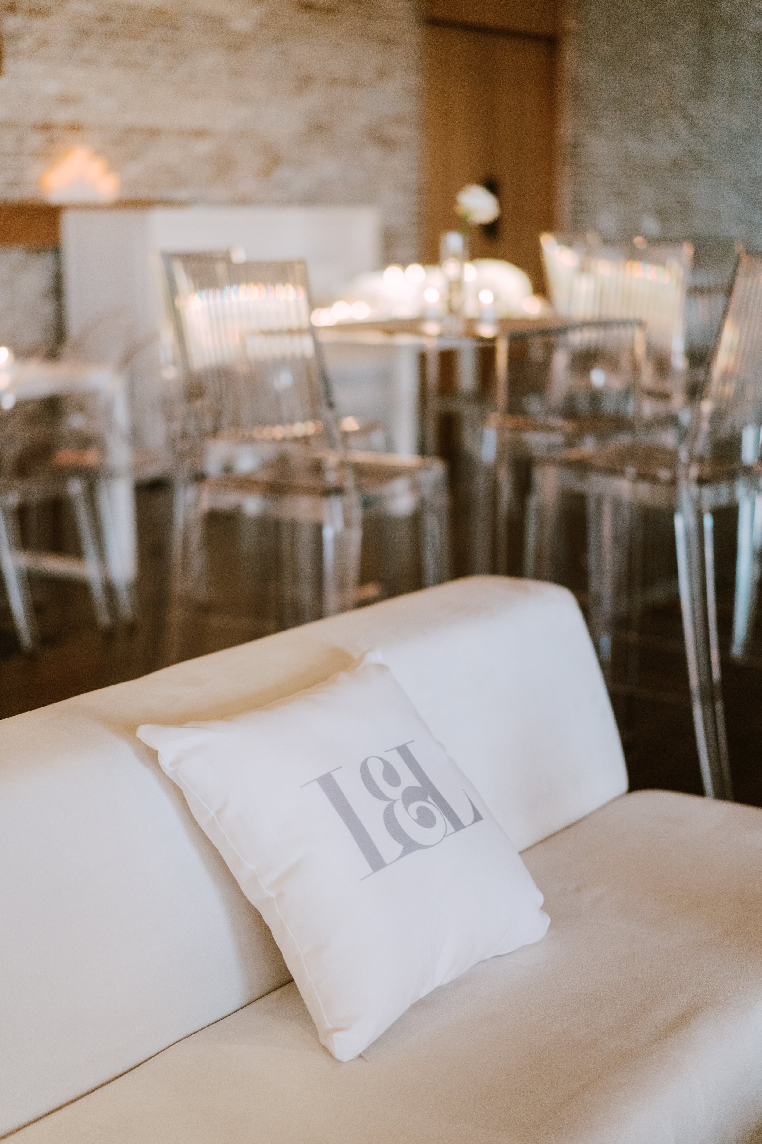 A white sofa with a pillow that has an engraved logo, in front of a dining area with a round table and clear acrylic chairs, in a warmly lit room with exposed brick wall.