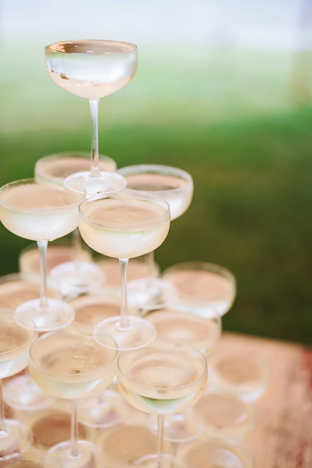 A pyramid of clear champagne glasses filled with a transparent, light-colored liquid, set against a blurred green outdoor background.