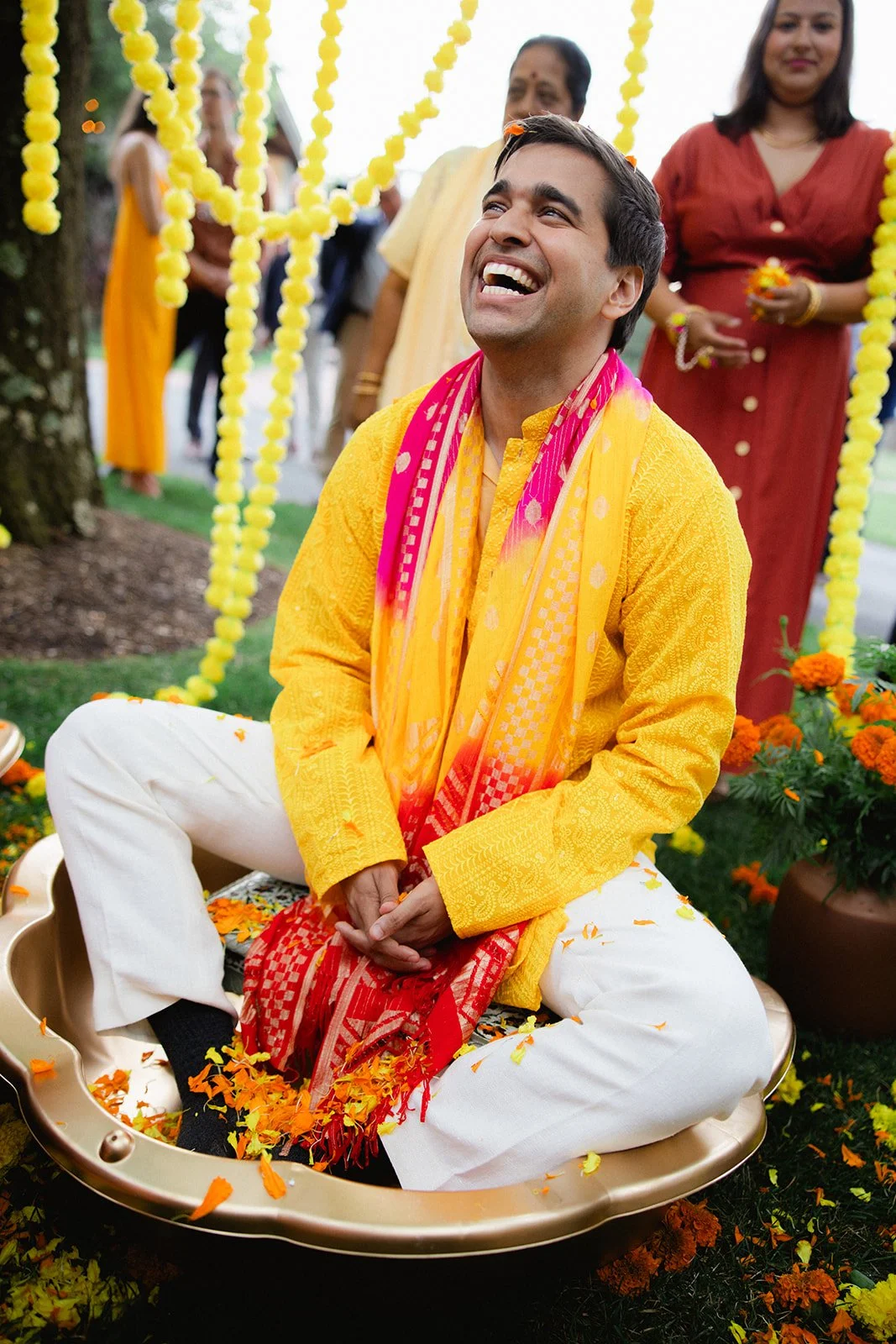 A joyful man in traditional Indian clothing sitting cross-legged on a large dish during a celebration, surrounded by women and decorated with yellow and orange flowers and marigolds.