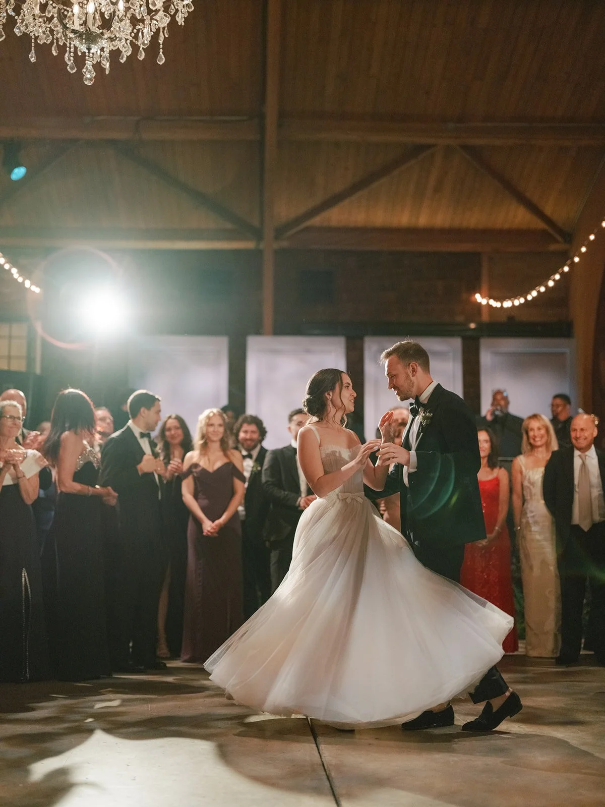 A bride and groom dancing at their wedding reception, surrounded by guests. The bride is wearing a white wedding gown and the groom is in a black tuxedo. The guests are watching and smiling.