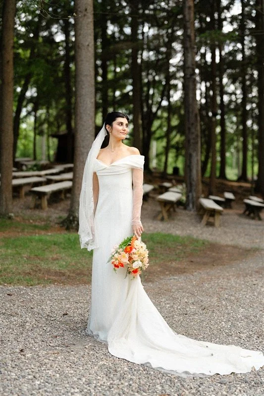 Bride in a white wedding dress holding a bouquet of orange and white flowers outdoors with trees and wooden benches in the background.