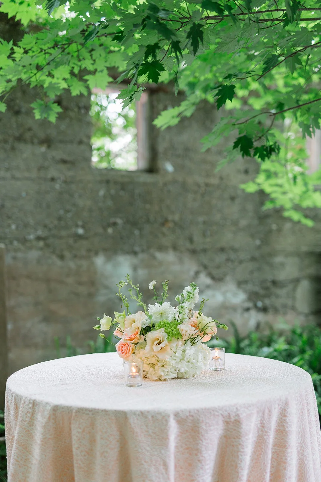 A round table with a pink lace tablecloth, decorated with a floral arrangement of white, peach, and green flowers, and two small lit candles in glass holders, set outdoors under leafy green trees.