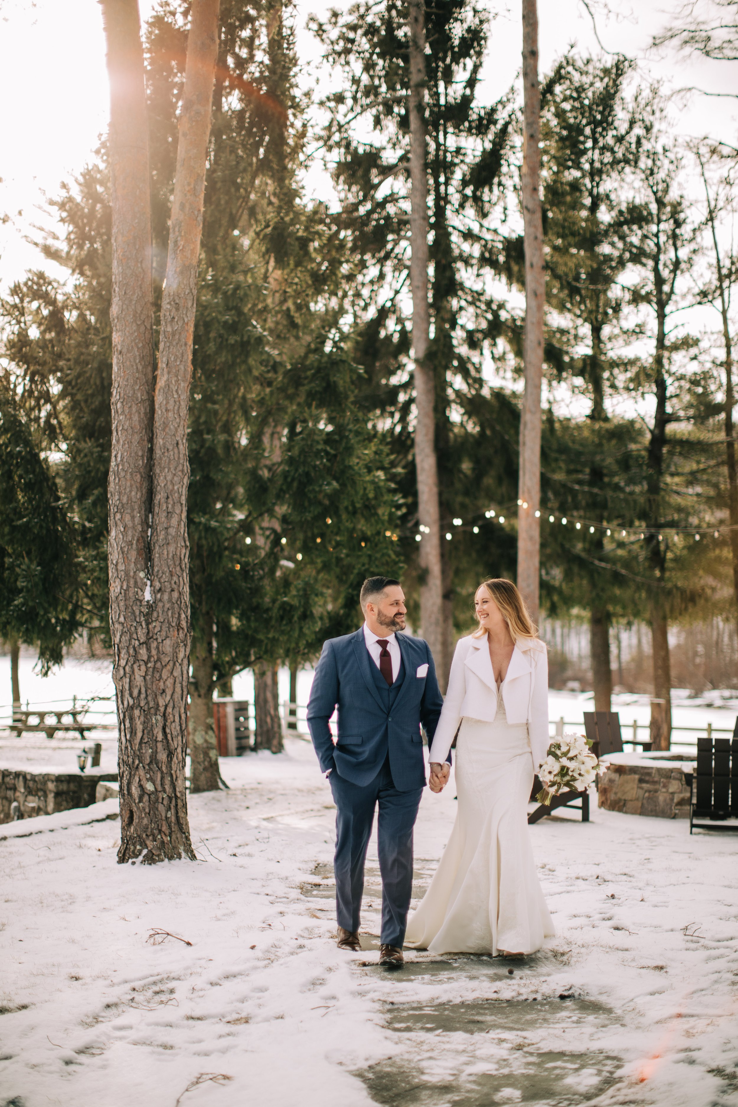 A newlywed couple walking hand in hand outdoors in a snowy forest, with string lights and trees in the background.
