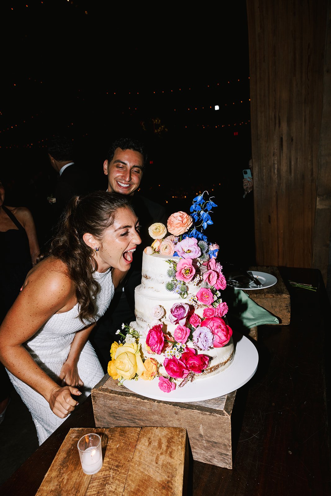 A woman and a man are celebrating a wedding with a large, multi-tiered white cake decorated with colorful flowers. The woman appears to be pretending to bite the cake while the man smiles in the background at a wedding reception.