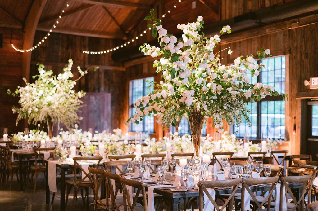 A rustic wedding reception with long tables decorated with large floral centerpieces of white flowers, wooden chairs, and string lights hanging from the wooden ceiling.