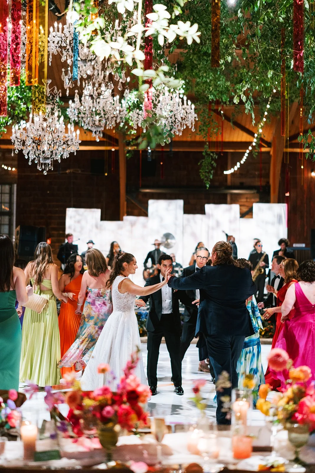 People dancing at a wedding reception with a band playing in the background, surrounded by colorful floral arrangements and hanging chandeliers.